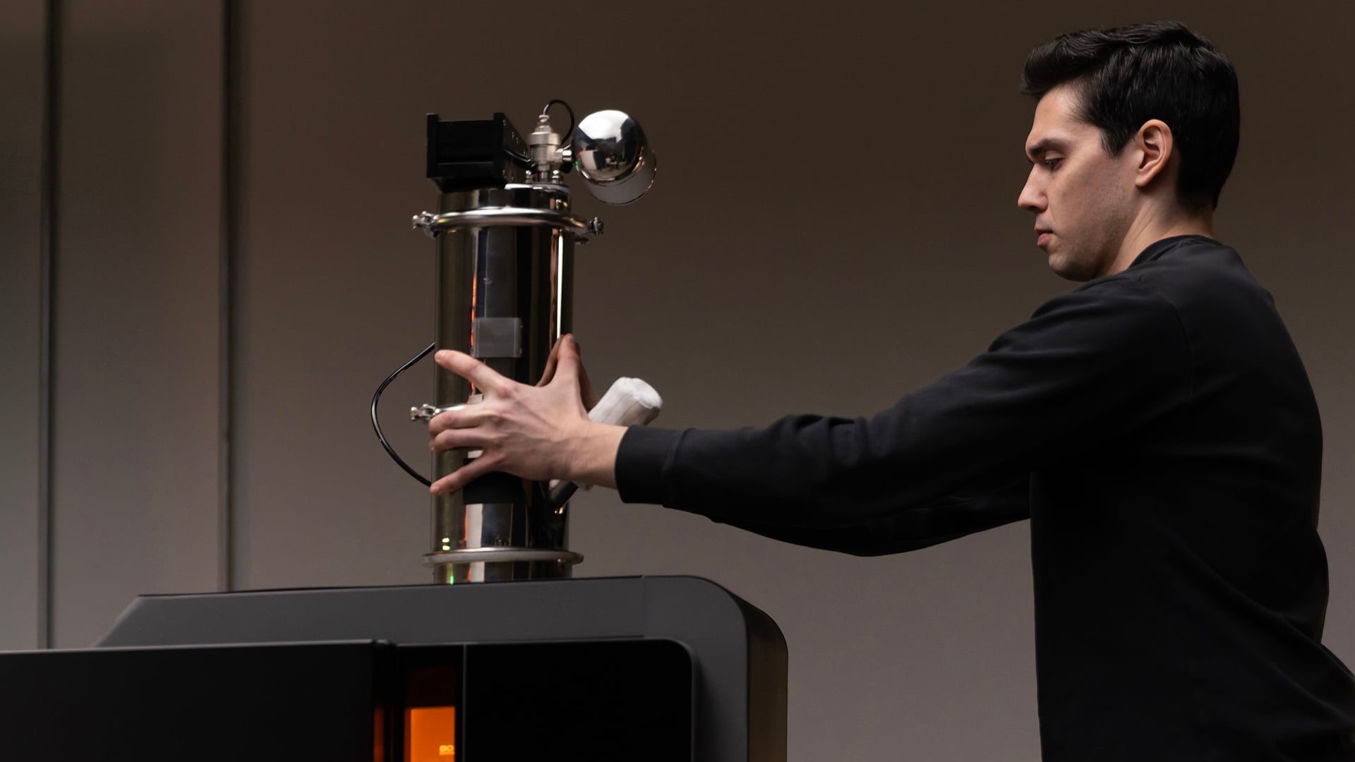 Technician in black long-sleeve shirt mounting the Formlabs Fuse X1 Vacuum Conveyor onto the top powder port of a Fuse X1 SLS printer; orange build-chamber glow visible through the front window