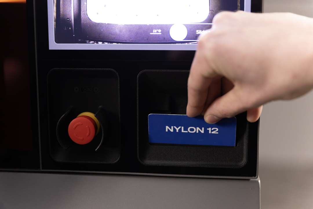 Close-up of a hand inserting a blue Nylon 12 NFC material card into the Formlabs Fuse X1 front-panel reader slot, red emergency stop button visible to the left